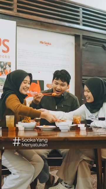Tiga orang duduk semeja saling menyuapkan lauk sambil tertawa di Mie Sooka